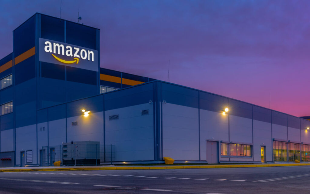 Image of the outside of an Amazon warehouse at dusk with the Amazon logo and a few external lights illuminated